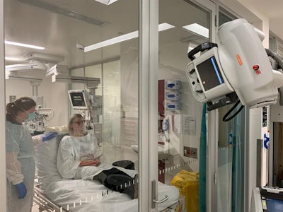 The radiographer inside the cubicle positions the bed close to the glass door and places the digital detector behind the patient for an erect anterior-posterior chest x-ray before stepping away from the patient during the exposure. The radiographer outside the cubicle positions the x-ray tube head close to the glass door and steps laterally away to initiate the x-ray while maintaining visual contact with the patient. The subject in the figure is not a patient but the author of this publication. Reproduced with permission of the Australasian College of Physical Scientists and Engineers in Medicine (ACPSEM).