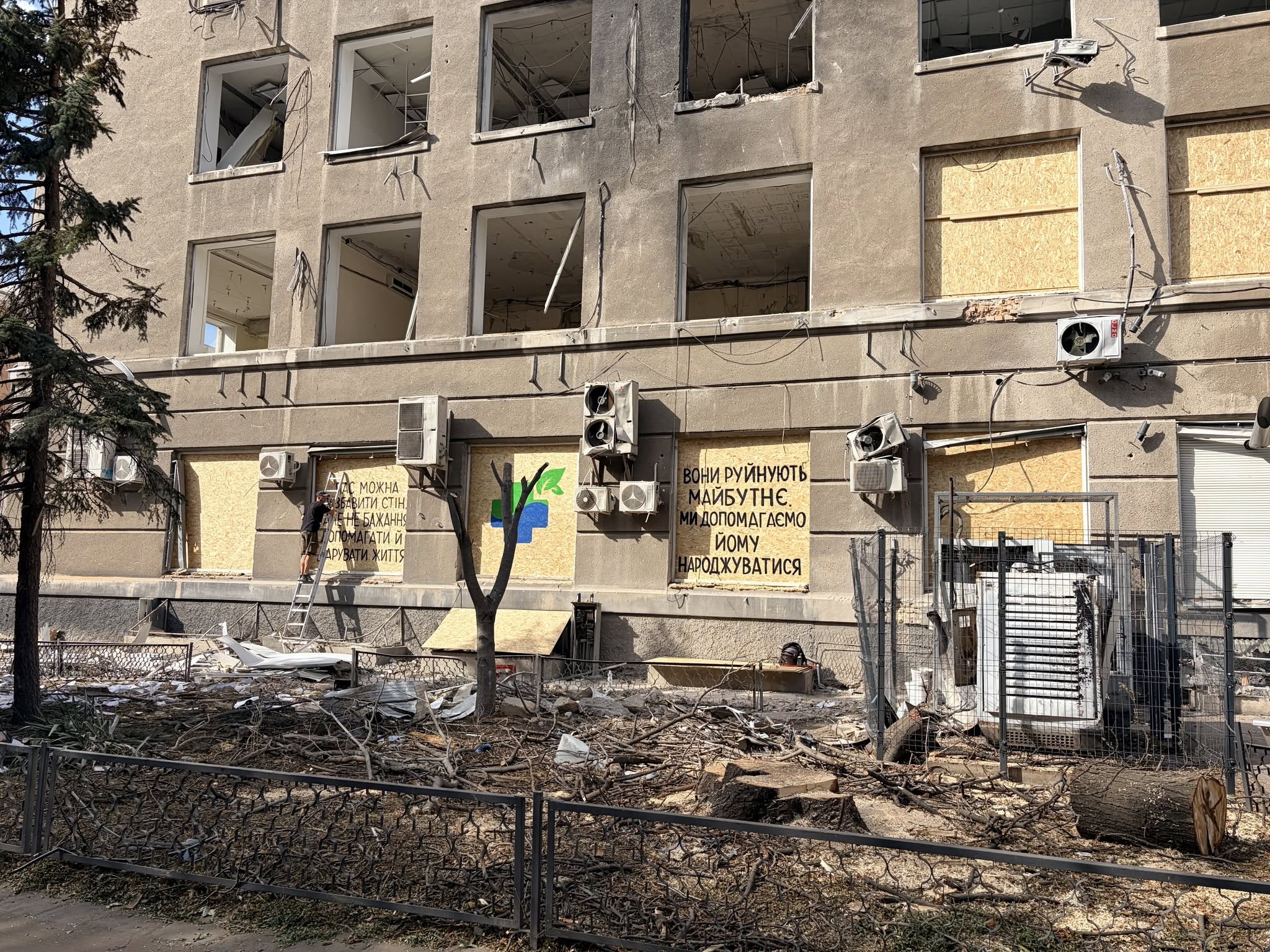 The exterior of Innova Diagnostica shows shattered windows, exposed wiring, damaged air-conditioning units, and debris surrounding the building. Several ground-floor windows have been boarded up. A message painted on the facade reads, “They destroy the future. We help bring it to life.”