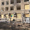 The exterior of Innova Diagnostica shows shattered windows, exposed wiring, damaged air-conditioning units, and debris surrounding the building. Several ground-floor windows have been boarded up. A message painted on the facade reads, “They destroy the future. We help bring it to life.”