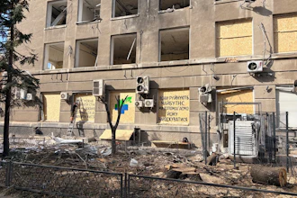 The exterior of Innova Diagnostica shows shattered windows, exposed wiring, damaged air-conditioning units, and debris surrounding the building. Several ground-floor windows have been boarded up. A message painted on the facade reads, “They destroy the future. We help bring it to life.”