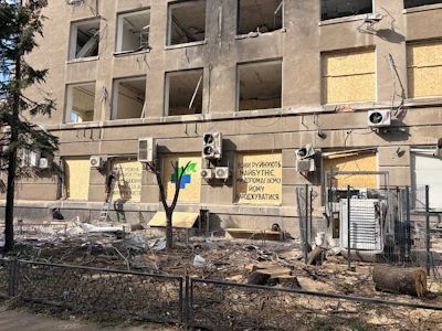 The exterior of Innova Diagnostica shows shattered windows, exposed wiring, damaged air-conditioning units, and debris surrounding the building. Several ground-floor windows have been boarded up. A message painted on the facade reads, “They destroy the future. We help bring it to life.”