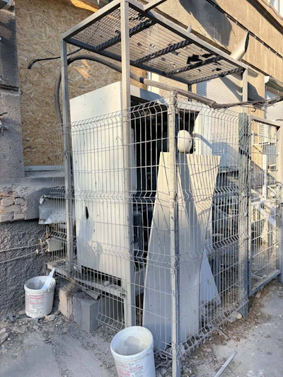An outdoor technical unit behind a protective metal fence shows visible damage to its casing and surrounding structure. The precise function of the equipment should be confirmed with Innova Diagnostica.