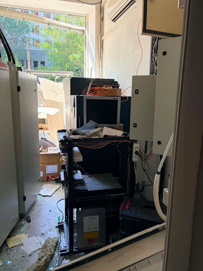 Broken glass and debris cover the floor of a technical room inside Innova Diagnostica. Equipment cabinets and an electronics rack appear exposed and displaced following damage to the building.
