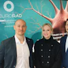 Three professionals posing at NeuroRad Next Generation conference event with turquoise branded backdrop featuring neural imagery
