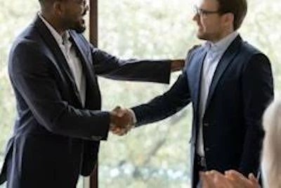Two businessmen in suits shaking hands during a professional meeting with a blurred window background