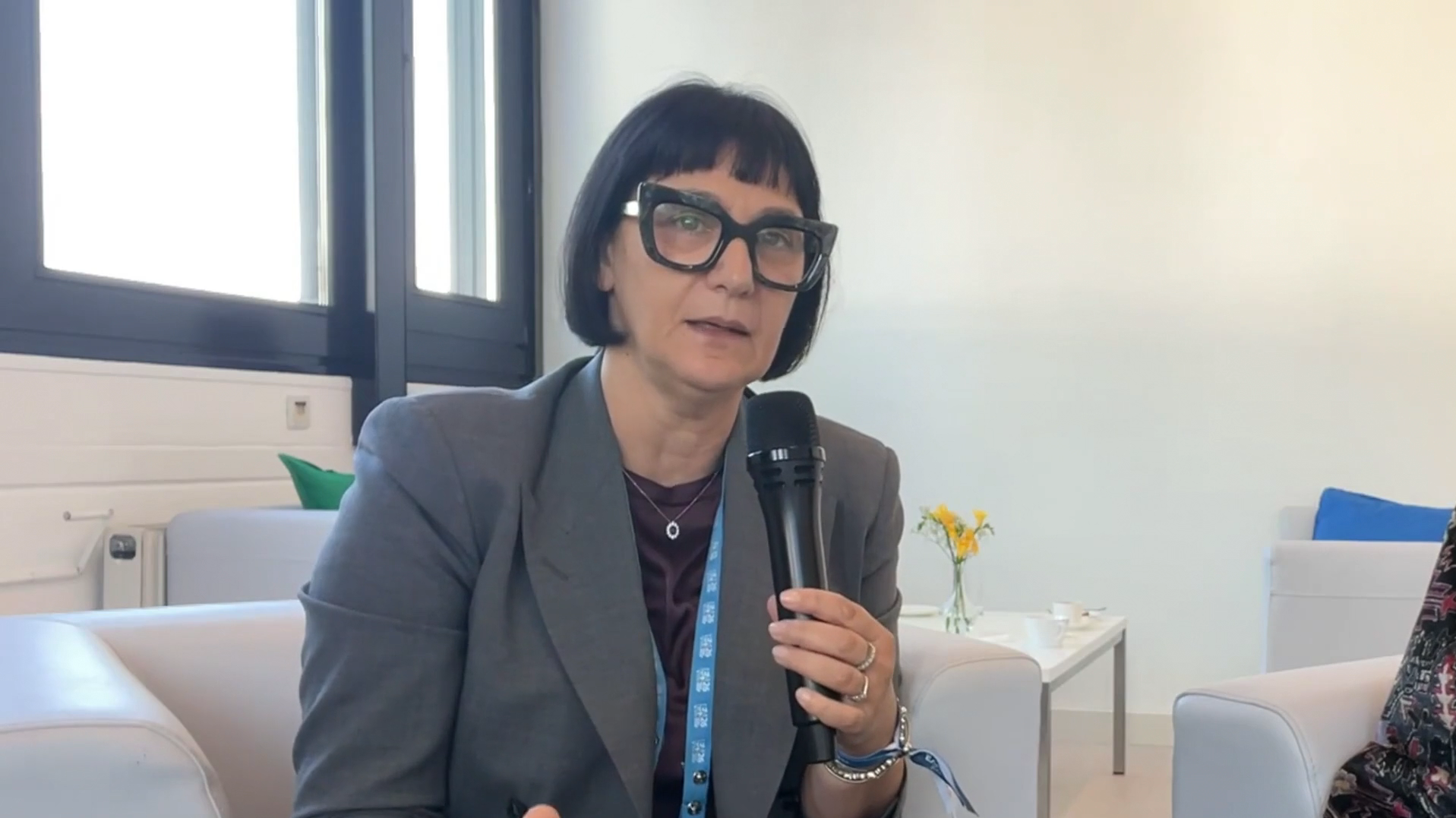 Woman wearing glasses and gray blazer speaking at a microphone during a professional conference or event