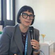 Woman wearing glasses and gray blazer speaking at a microphone during a professional conference or event