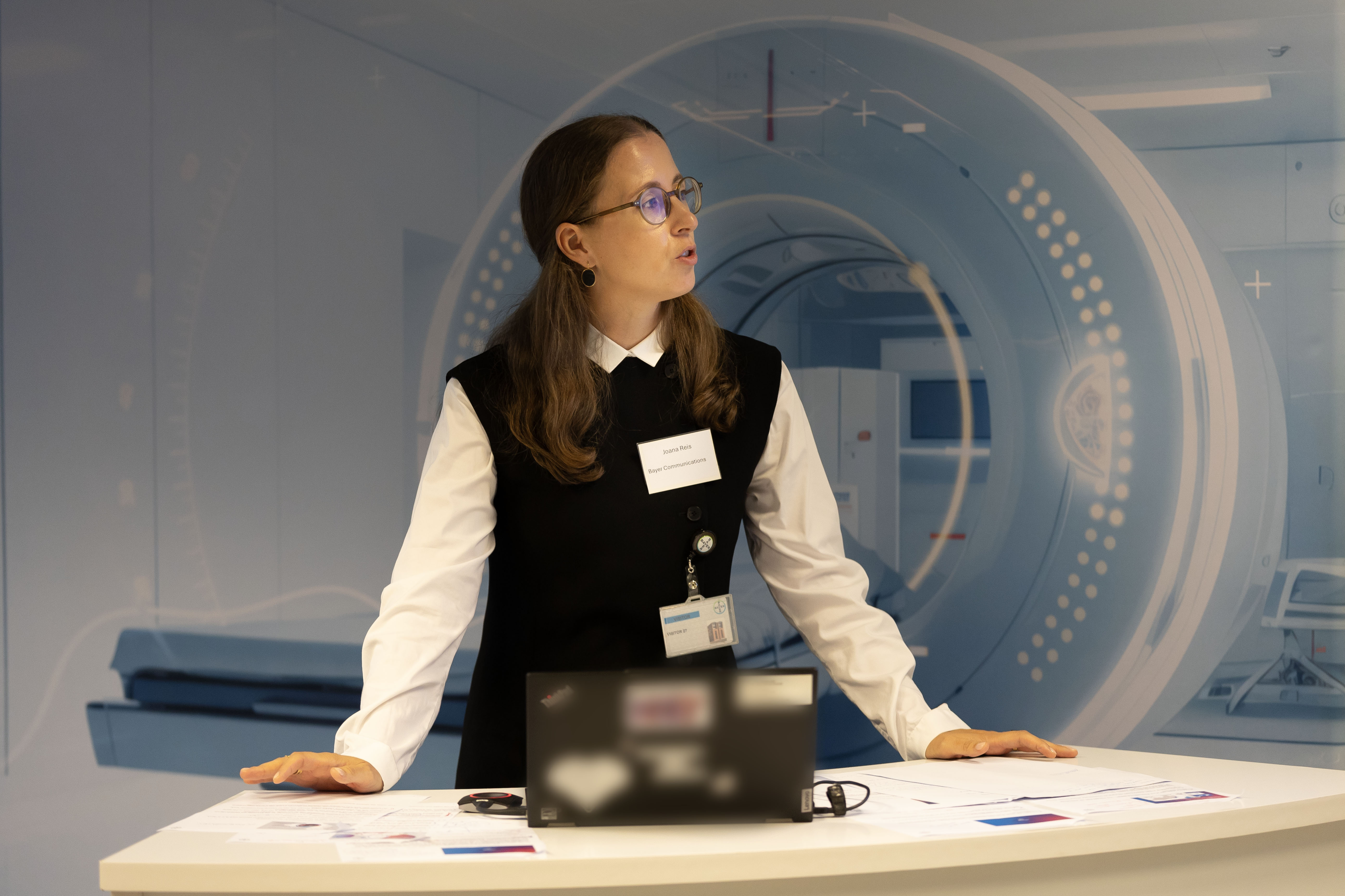 Professional woman at reception desk wearing glasses and name badge, with abstract blue geometric design on wall behind her