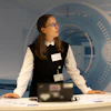 Professional woman at reception desk wearing glasses and name badge, with abstract blue geometric design on wall behind her