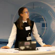 Professional woman at reception desk wearing glasses and name badge, with abstract blue geometric design on wall behind her