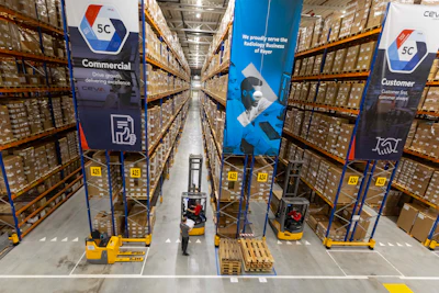 Automated warehouse aisle with yellow pallet jacks and 5C CEVA logistics branded banners on tall shelving units