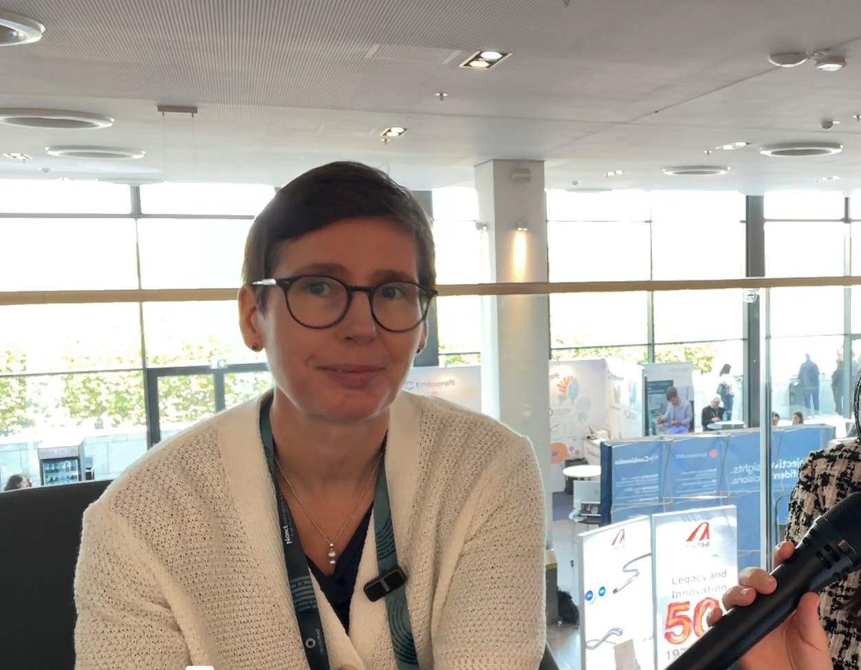 Professional woman wearing glasses and beige cardigan at indoor conference venue with modern architecture and blue displays