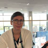 Professional woman wearing glasses and beige cardigan at indoor conference venue with modern architecture and blue displays