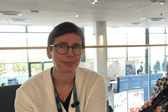 Professional woman wearing glasses and beige cardigan at indoor conference venue with modern architecture and blue displays