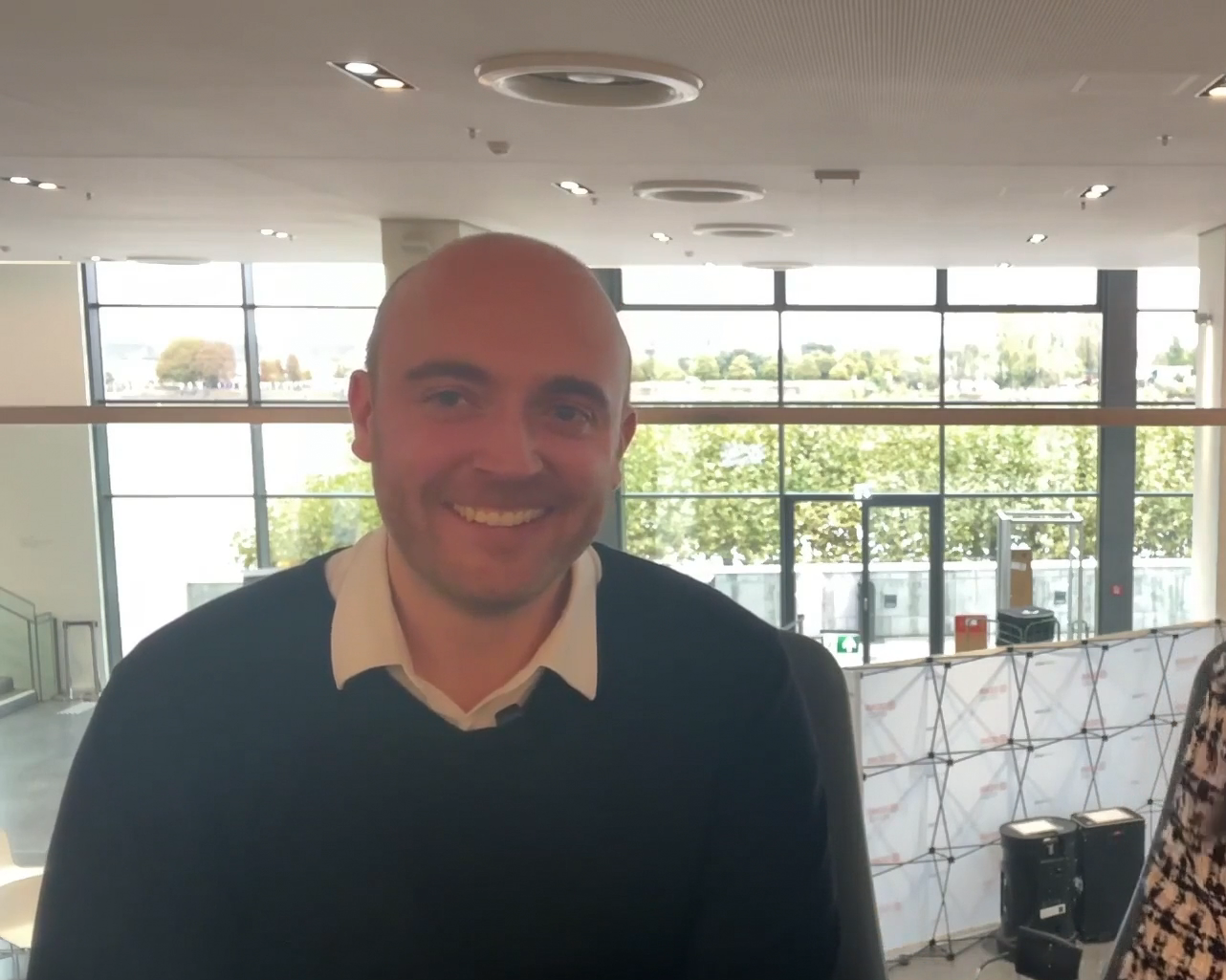 Smiling man in dark sweater with cream collar standing in modern office with large windows overlooking green landscape