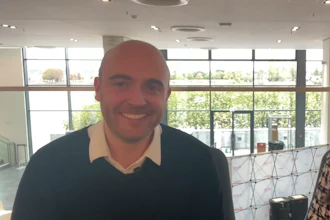Smiling man in dark sweater with cream collar standing in modern office with large windows overlooking green landscape
