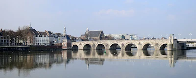 Historic stone bridge with multiple arches spanning calm river, European cityscape with church spires in background