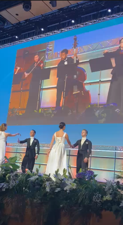 Formal event on stage with elegantly dressed guests and musicians performing on large screen display above