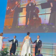 Formal event on stage with elegantly dressed guests and musicians performing on large screen display above