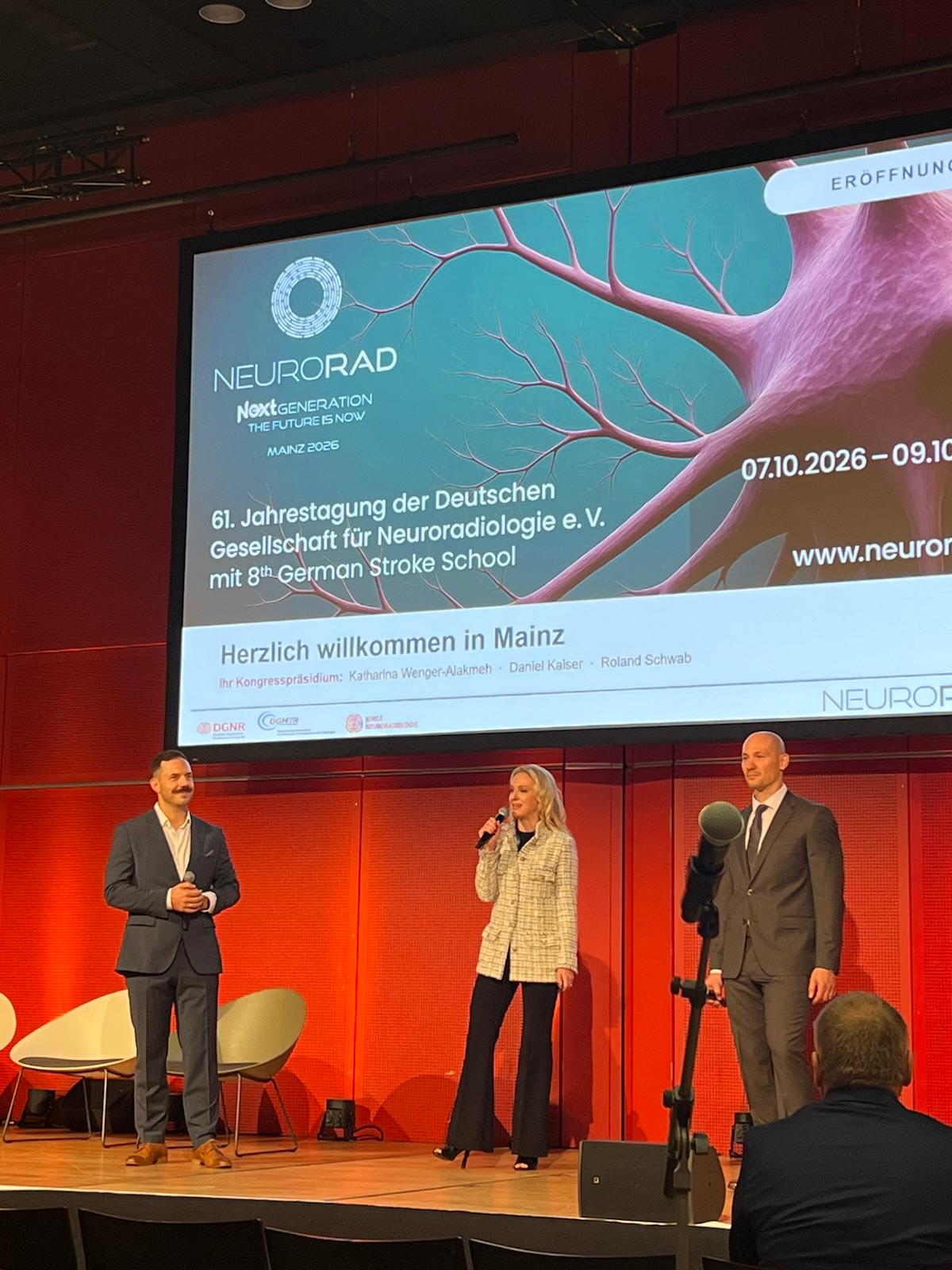 Three speakers presenting at NEURORAD 2026 conference in Mainz, with neurological imagery displayed on screen behind them