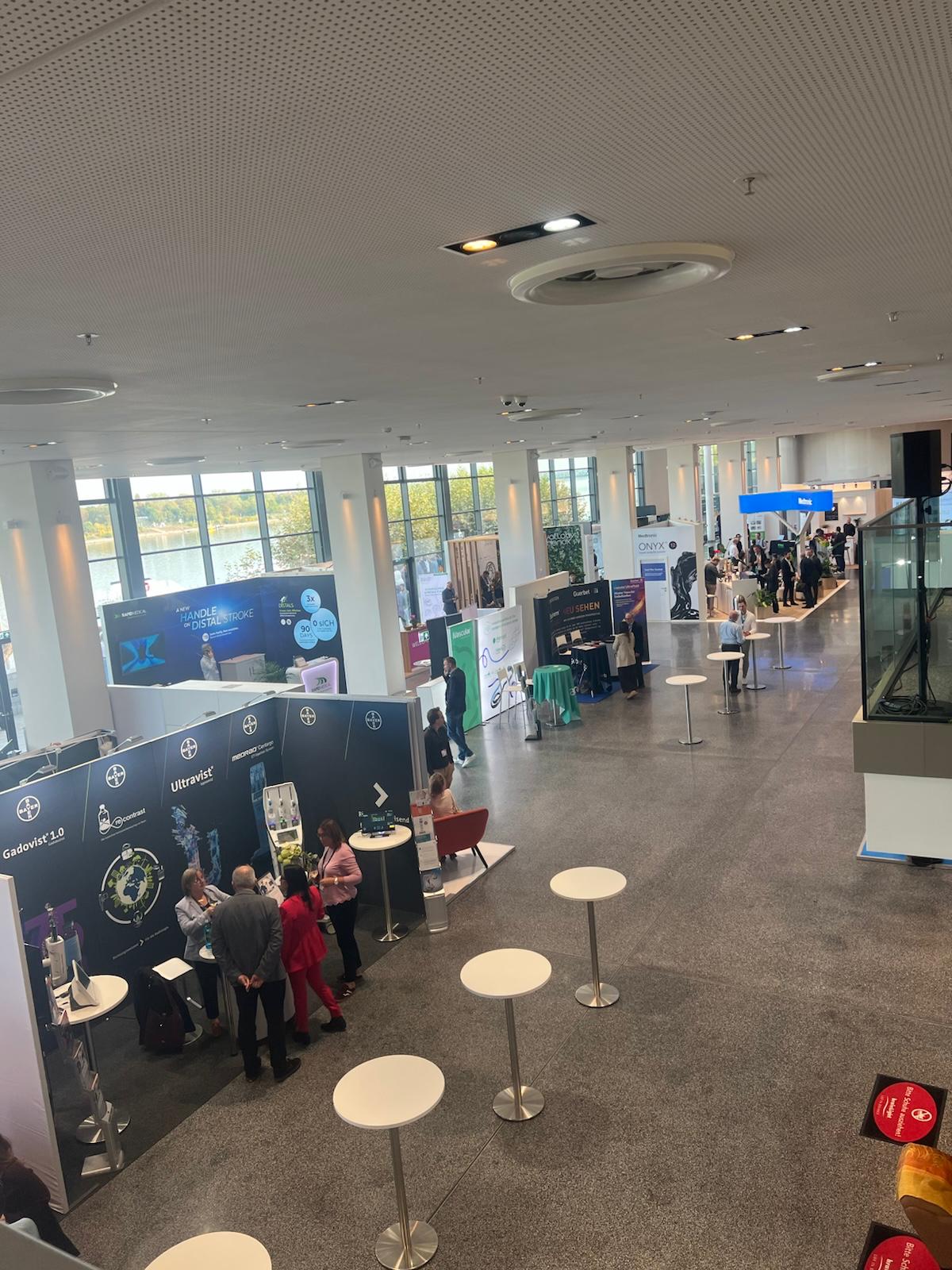 Modern conference hall with multiple branded exhibition booths, standing tables, and attendees networking during a corporate event.