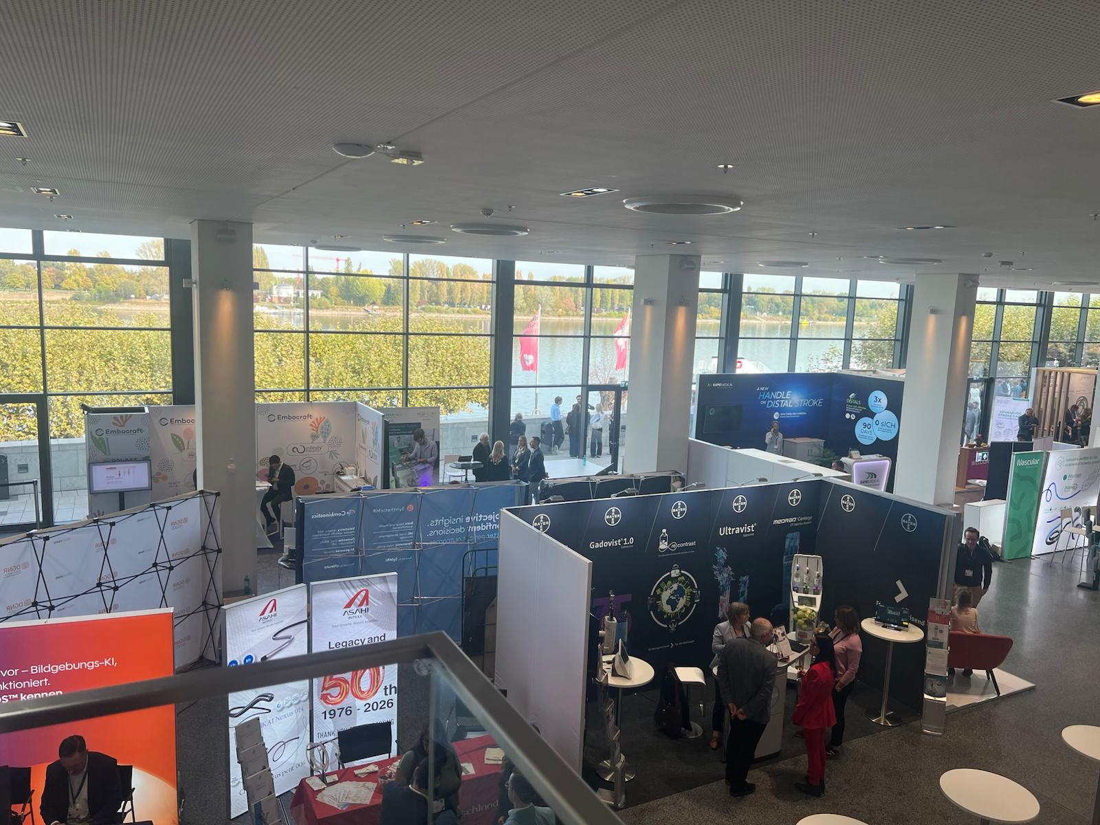 Modern exhibition hall with multiple branded booths, large windows overlooking autumn landscape, visitors exploring displays
