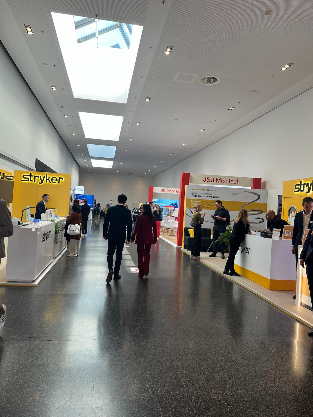 Trade show exhibition floor with Stryker and J&J MedTech branded booths, attendees in business attire, skylights overhead