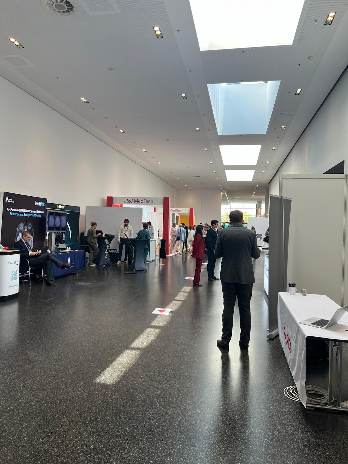 Professional trade show exhibition floor with multiple company booths, attendees in business attire, and modern ceiling with skylights