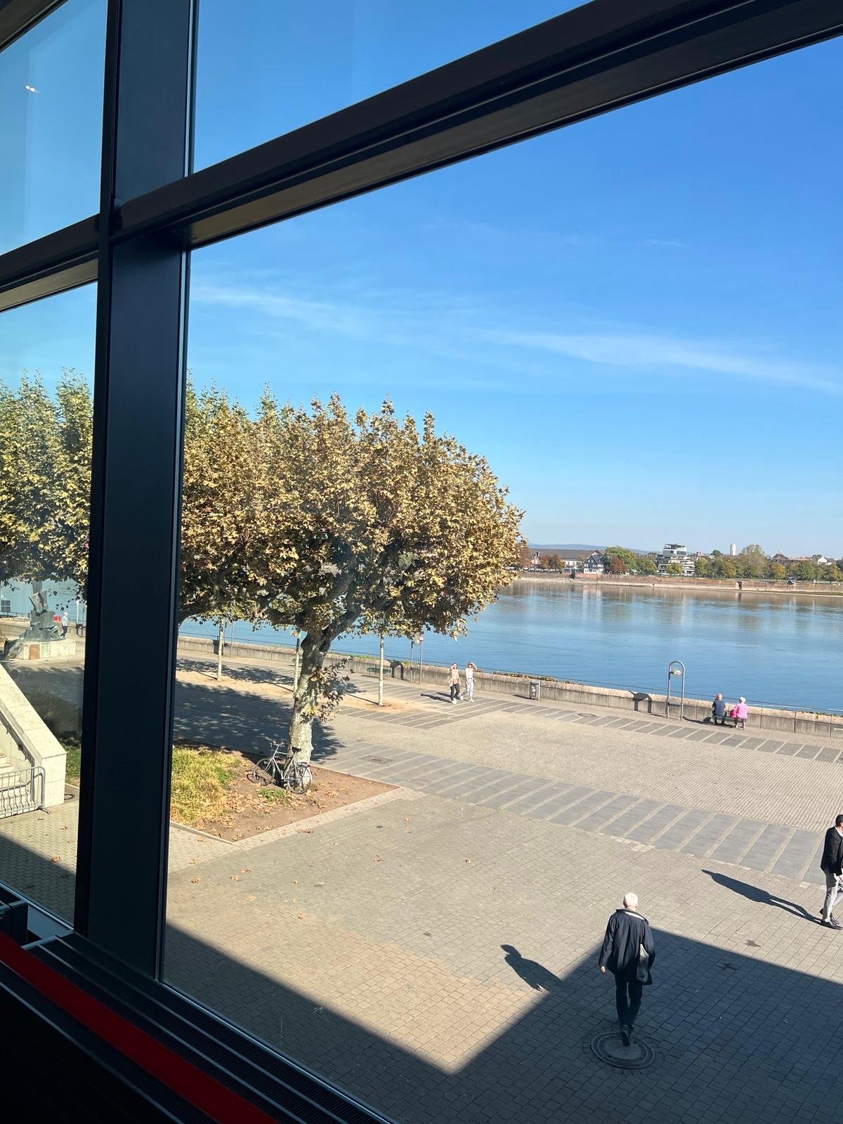 Riverside promenade view through large windows with tree, people, and calm blue water under clear sky