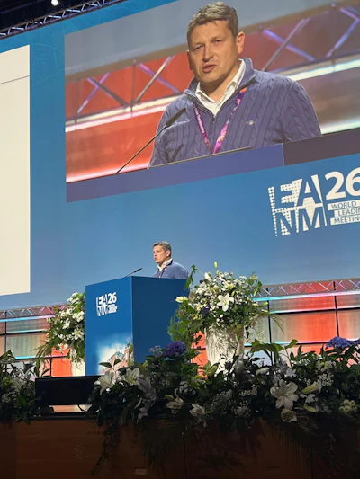 Professional speaker presenting at EA26 NIM World Leadership Meeting conference podium with floral arrangements