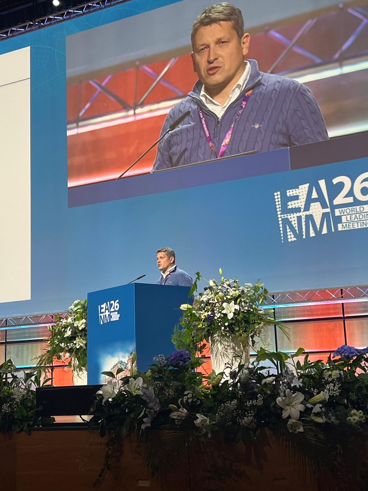 Speaker presenting at EA26 NIM World Leadership Meeting conference podium with floral arrangements