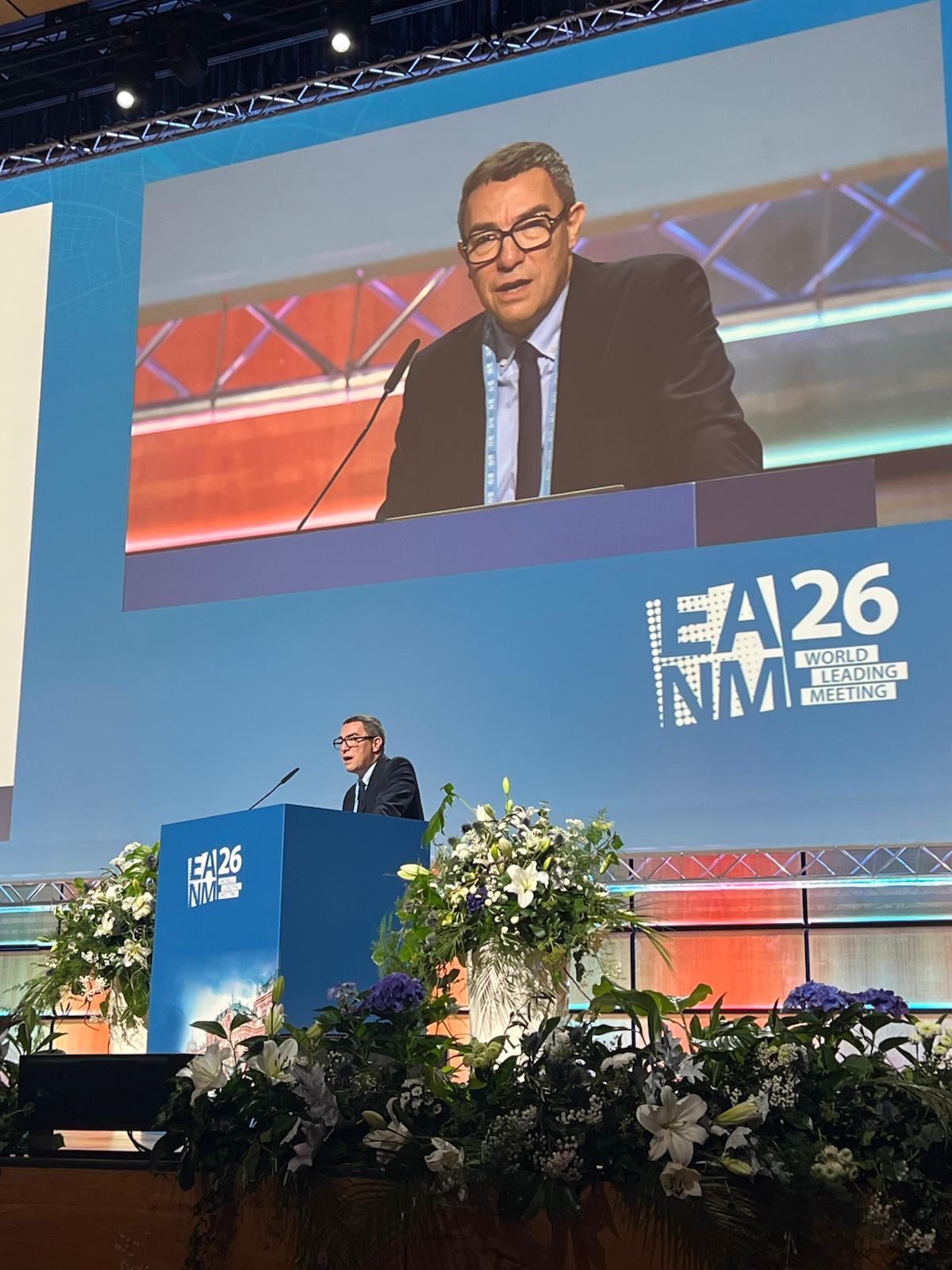 Speaker at EA26 NM conference podium with white flowers and large blue backdrop displaying event branding