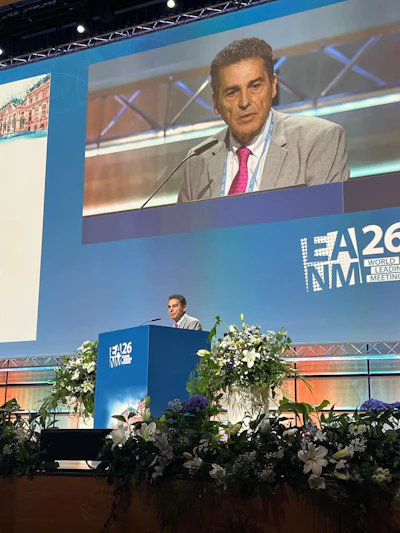 Speaker presenting at IEA26 World Leading Meeting conference podium with blue branding and floral arrangements