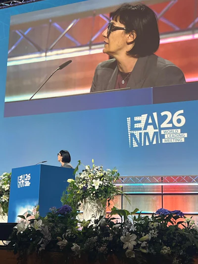 Speaker presenting at A26 NMI World Leading Meeting conference podium with large screen display