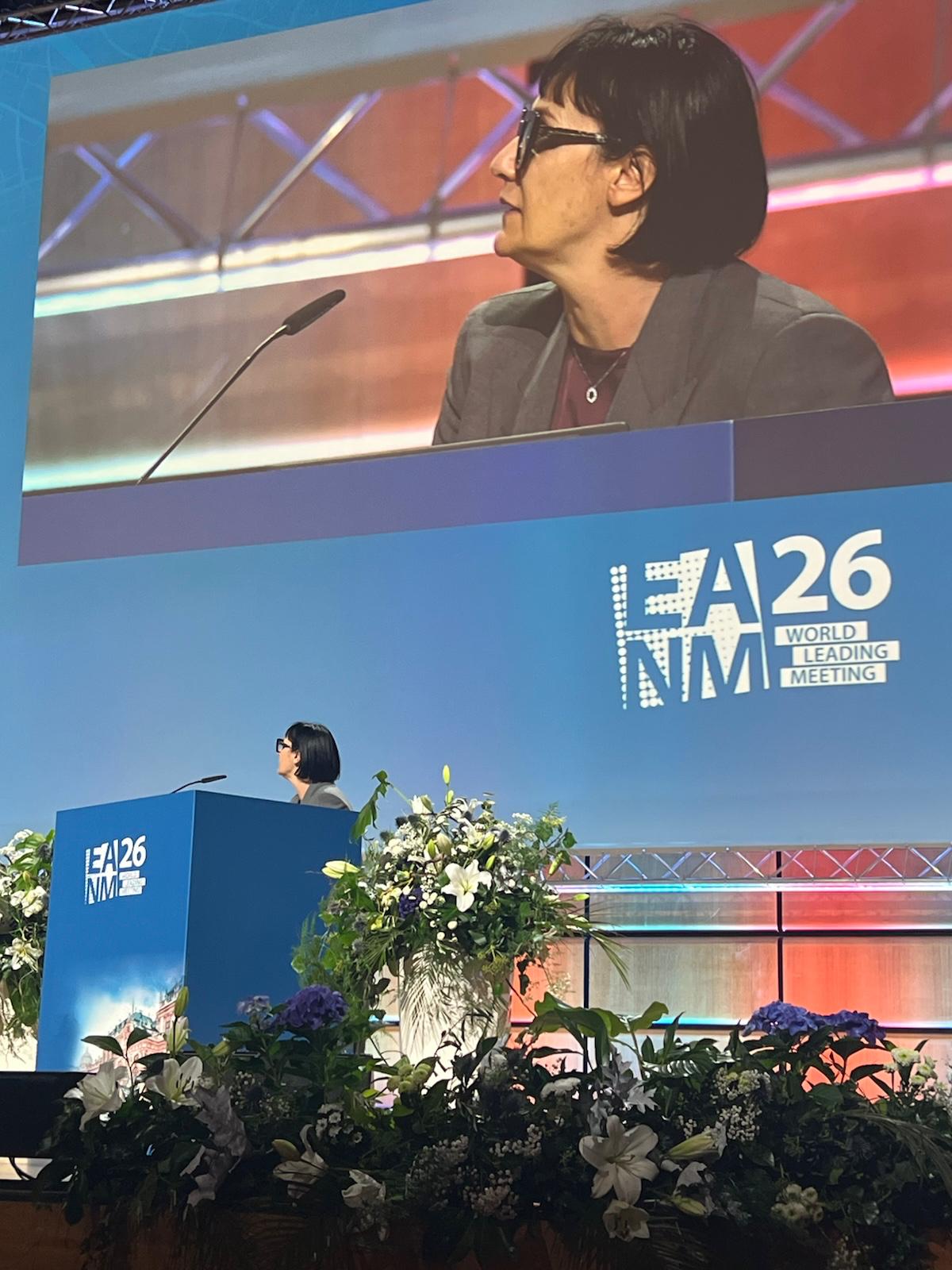 Speaker presenting at A26 NMA World Leading Meeting conference podium with floral arrangements