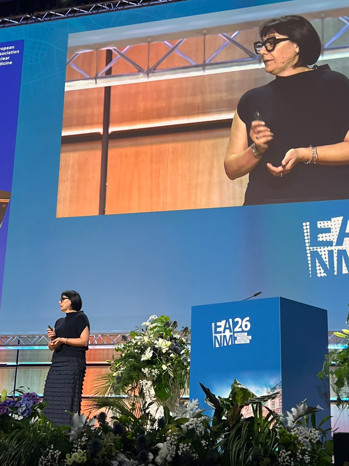 Female speaker presenting at EA26 NM conference with podium and large screen display