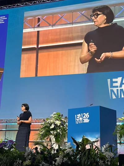 Woman speaker presenting at EA26 NM conference with large screen display and blue podium