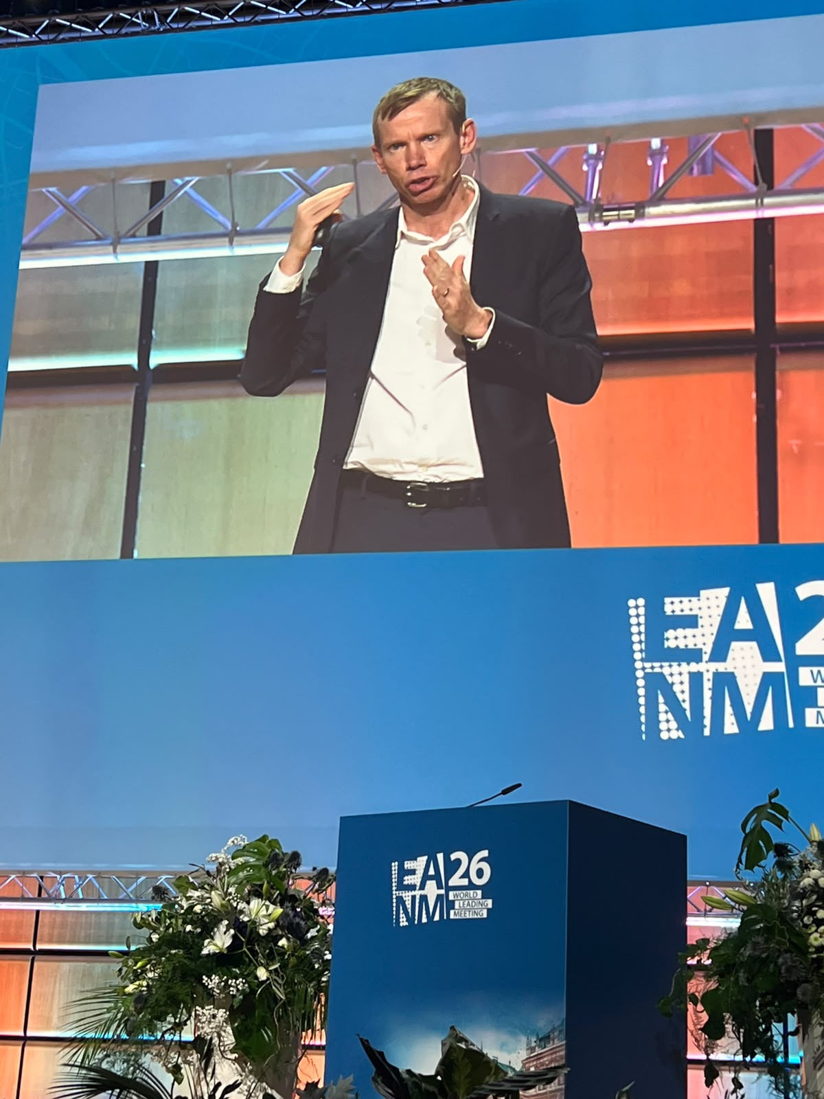 Man in dark blazer speaking at NME Awards event podium with blue and orange branded backdrop