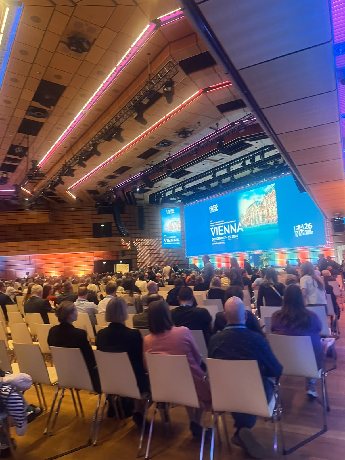 Conference hall with attendees seated facing a large blue projection screen displaying Vienna event branding and architectural imagery