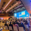 Conference hall with attendees seated facing a large blue projection screen displaying Vienna event branding and architectural imagery