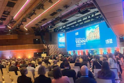 Conference hall with attendees seated facing a large blue projection screen displaying Vienna event branding and architectural imagery