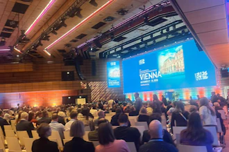 Conference hall with attendees seated facing a large blue projection screen displaying Vienna event branding and architectural imagery