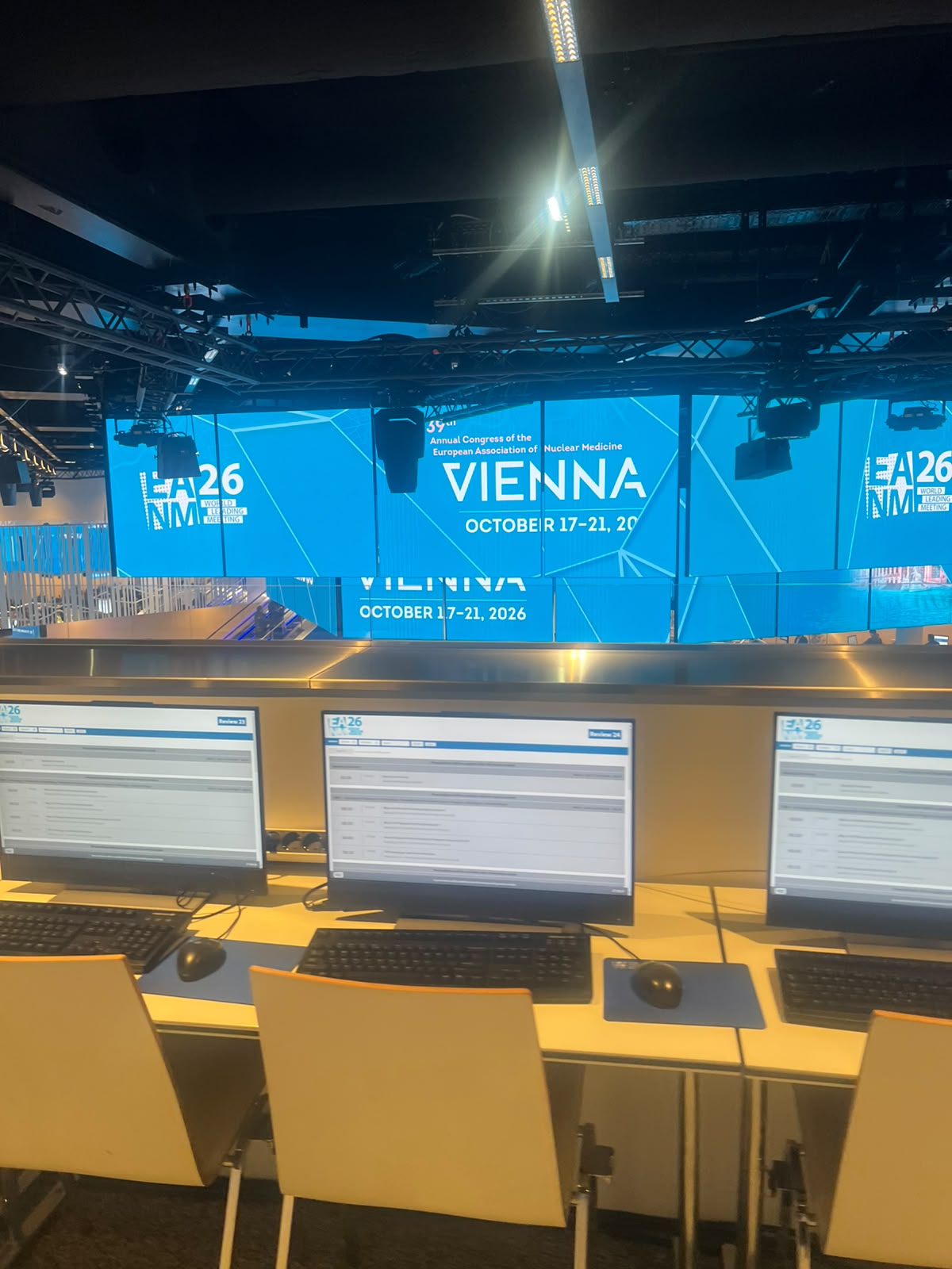 Conference venue setup with three computer workstations and yellow chairs facing a large blue event banner for HLM26 Vienna conference October 17-21, 2026