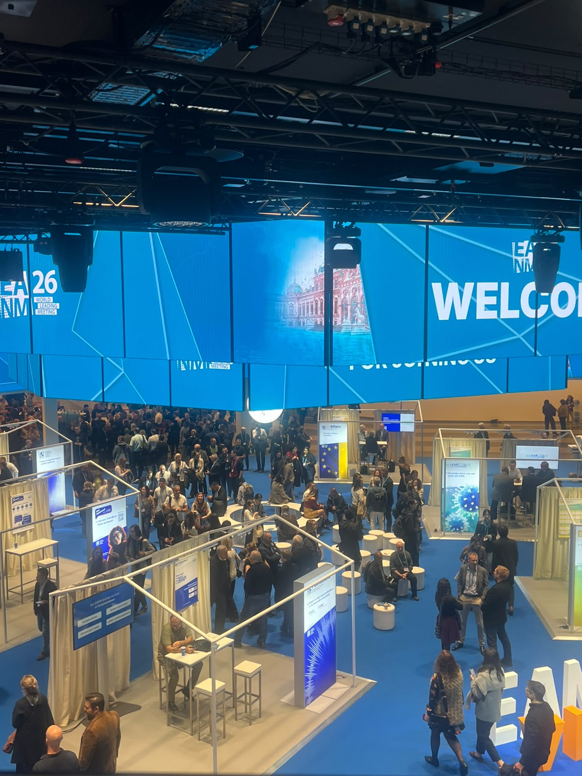 Crowded exhibition hall with blue-branded booths, overhead screens displaying welcome signage, and attendees navigating multiple wooden exhibition stands.