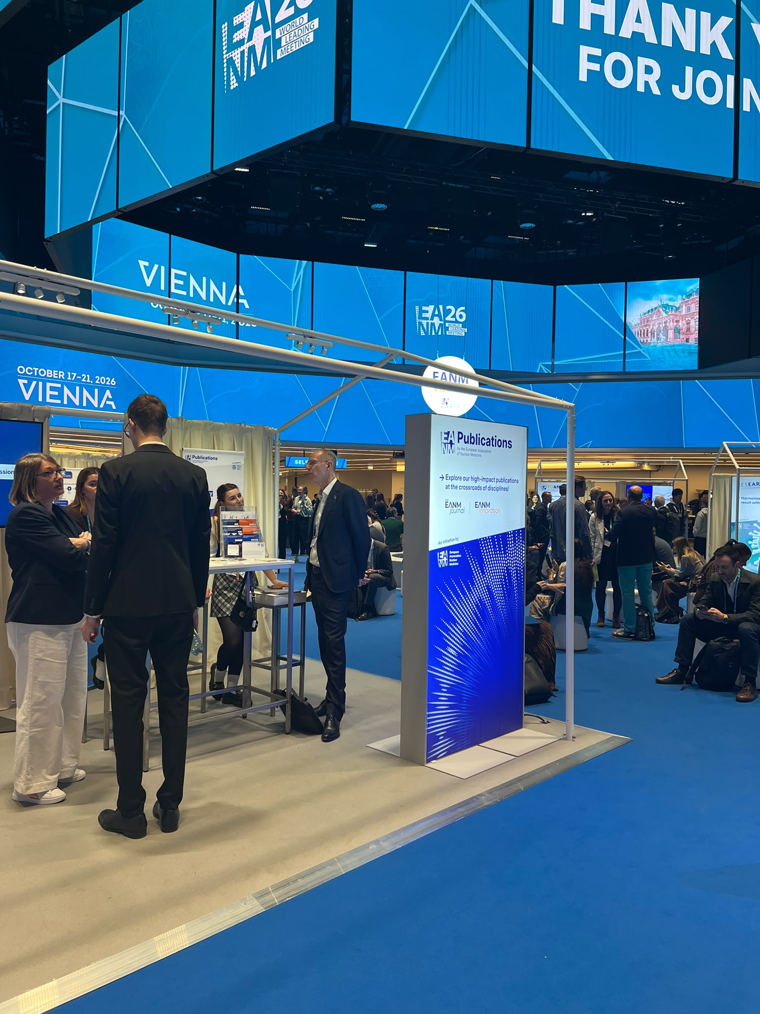 Professional conference booth at EA26 Vienna event with attendees networking, blue geometric design, and publications display.
