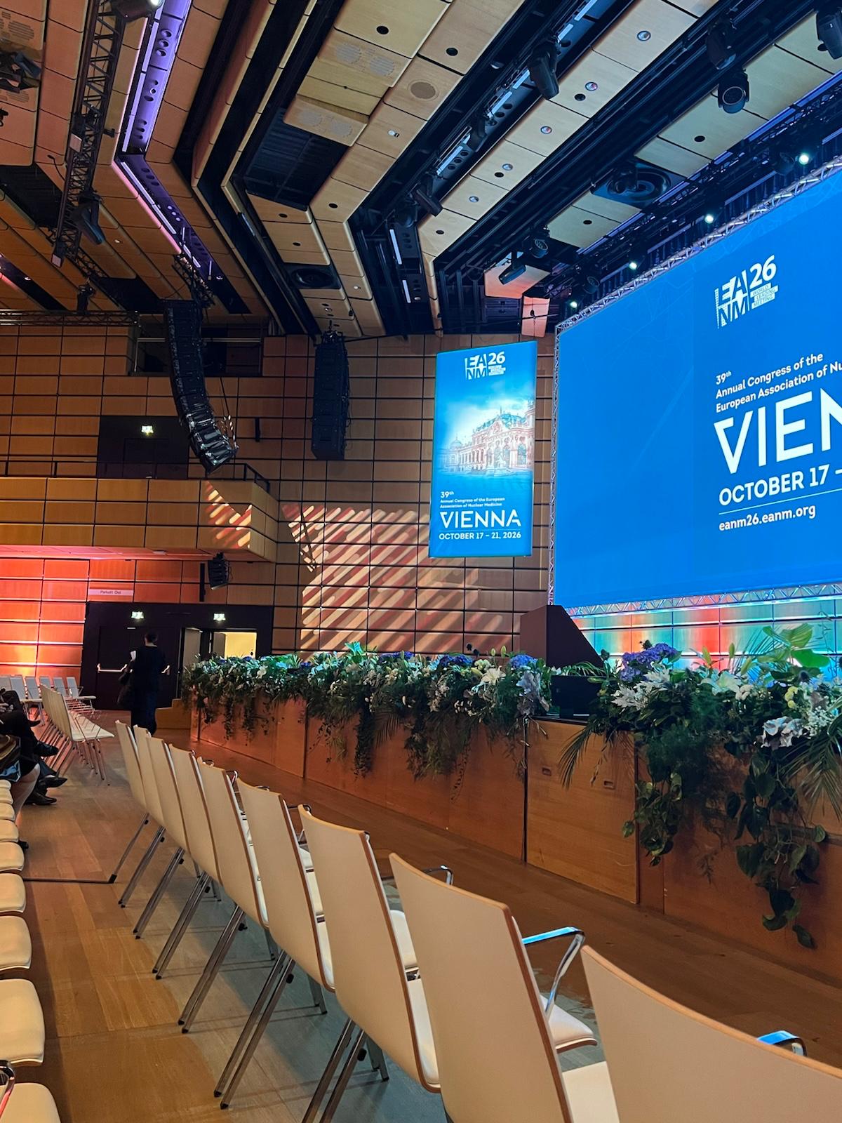 Conference venue with blue EAMM26 Vienna event signage displayed on large screens above seating area