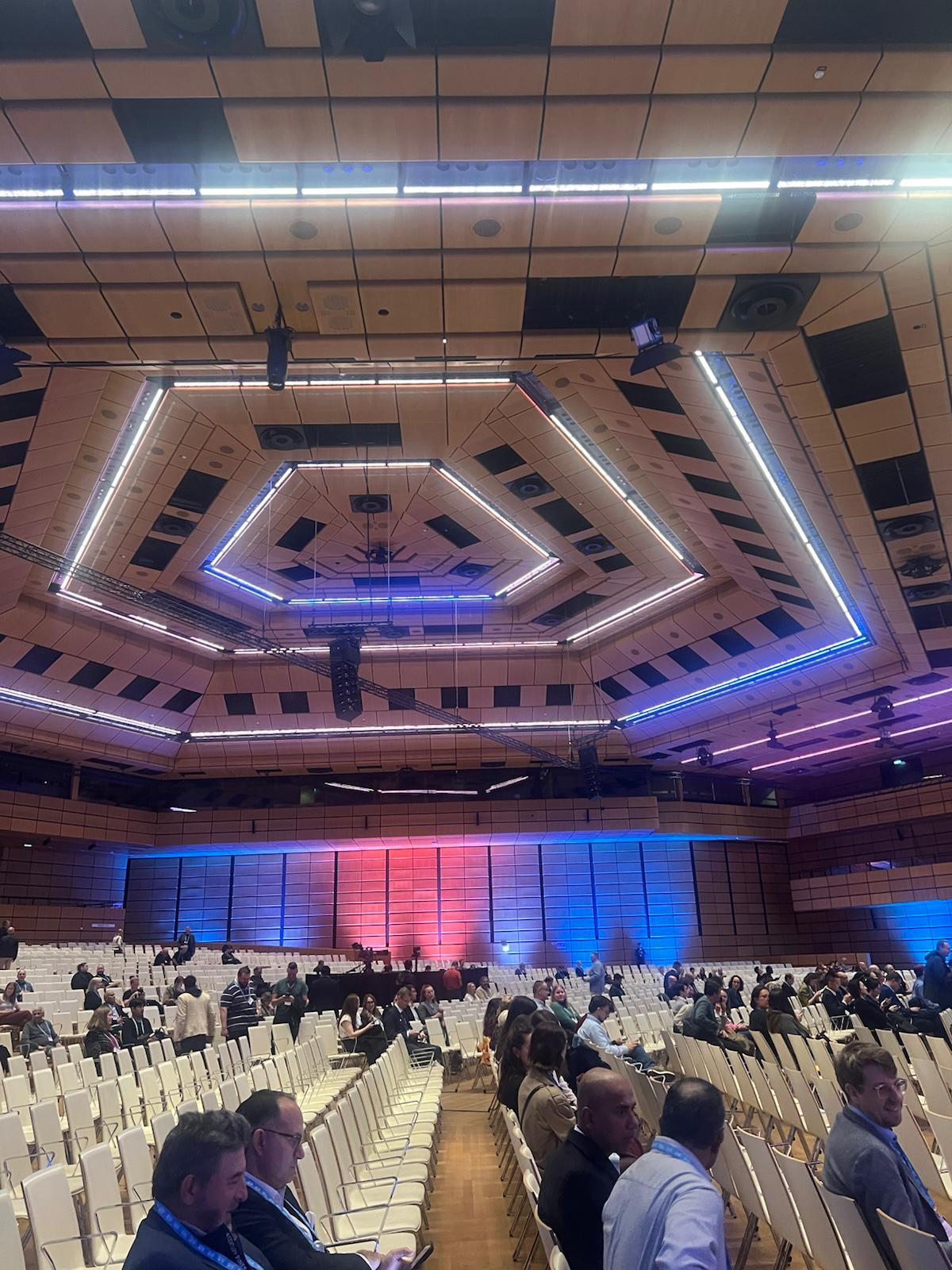 Modern auditorium interior with geometric neon-lit ceiling, tiered seating, and attendees at a conference or event venue