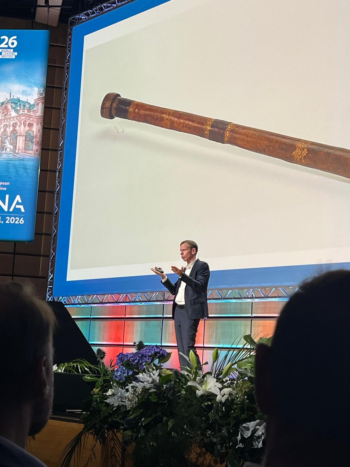 Professional speaker presenting at conference with historical artifact displayed on large screen behind podium