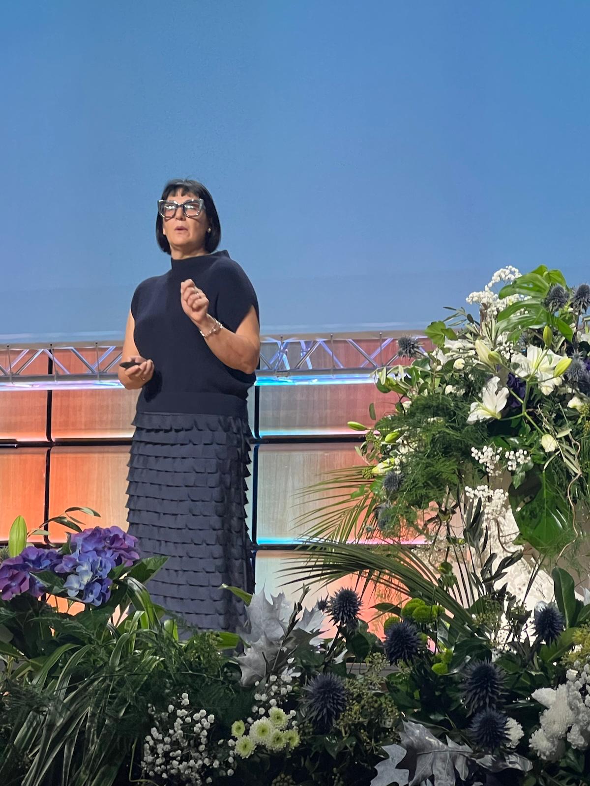 Woman speaking outdoors with decorative flowers and brick building backdrop under clear blue sky