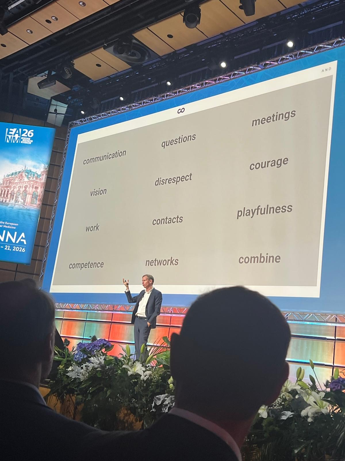 Speaker presenting at conference with word cloud display showing concepts like communication, vision, courage, and networks on large projection screen